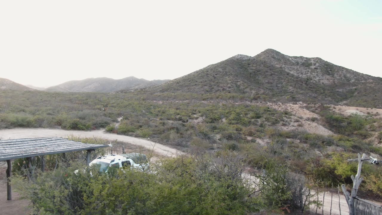 Cabo Pulmo desert view with a dirt road, partial carport, white SUV, green shrubs, distant rocky mountains, pale sky, and no visible people.