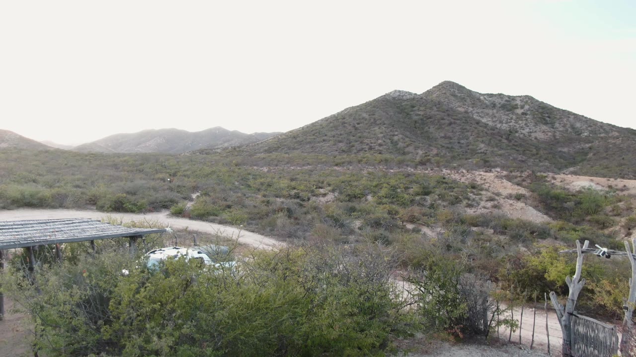 Cabo Pulmo frame with the carport and white SUV still visible near the dirt road, surrounded by scrub vegetation and backed by low mountains, with no visible people.