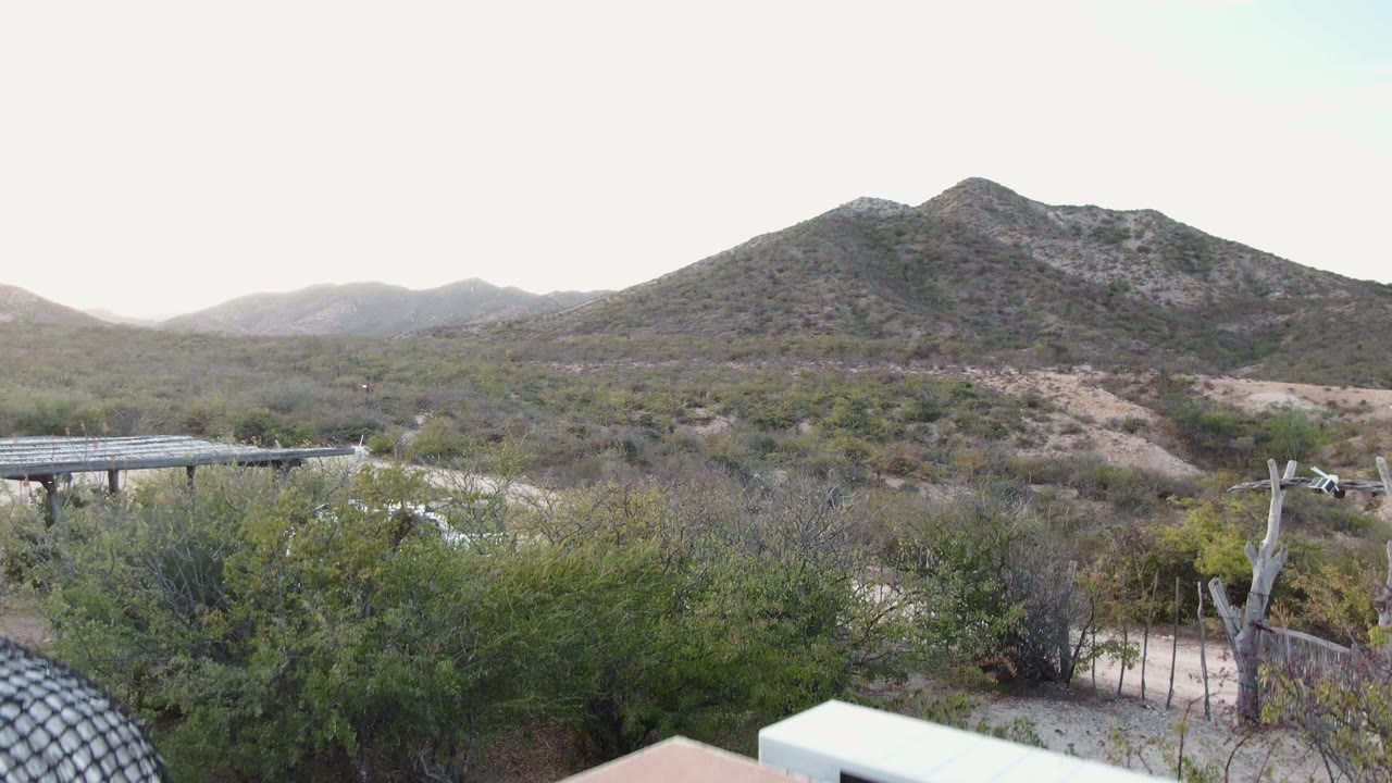 Mountain landscape with shrubs, road, fence, and roof edge entering foreground