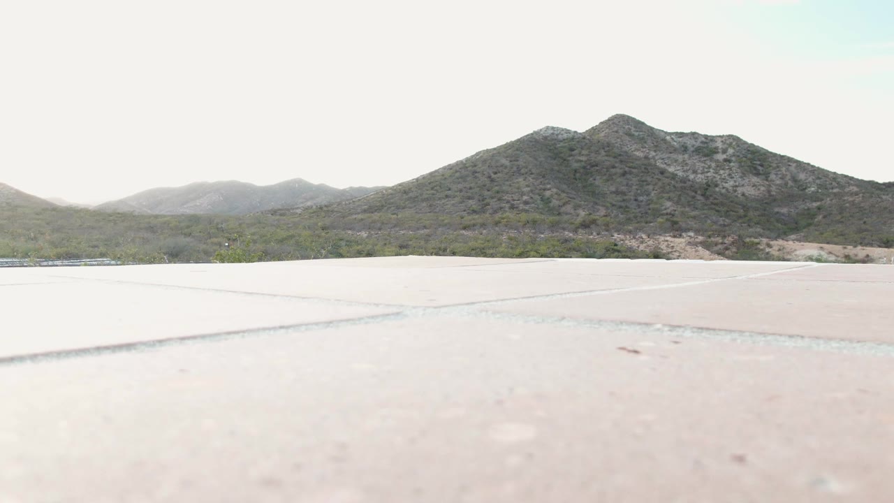 Cabo Pulmo, Baja California Sur frame showing a pale concrete area in front of low desert vegetation and a distant mountain ridge, with no visible people.