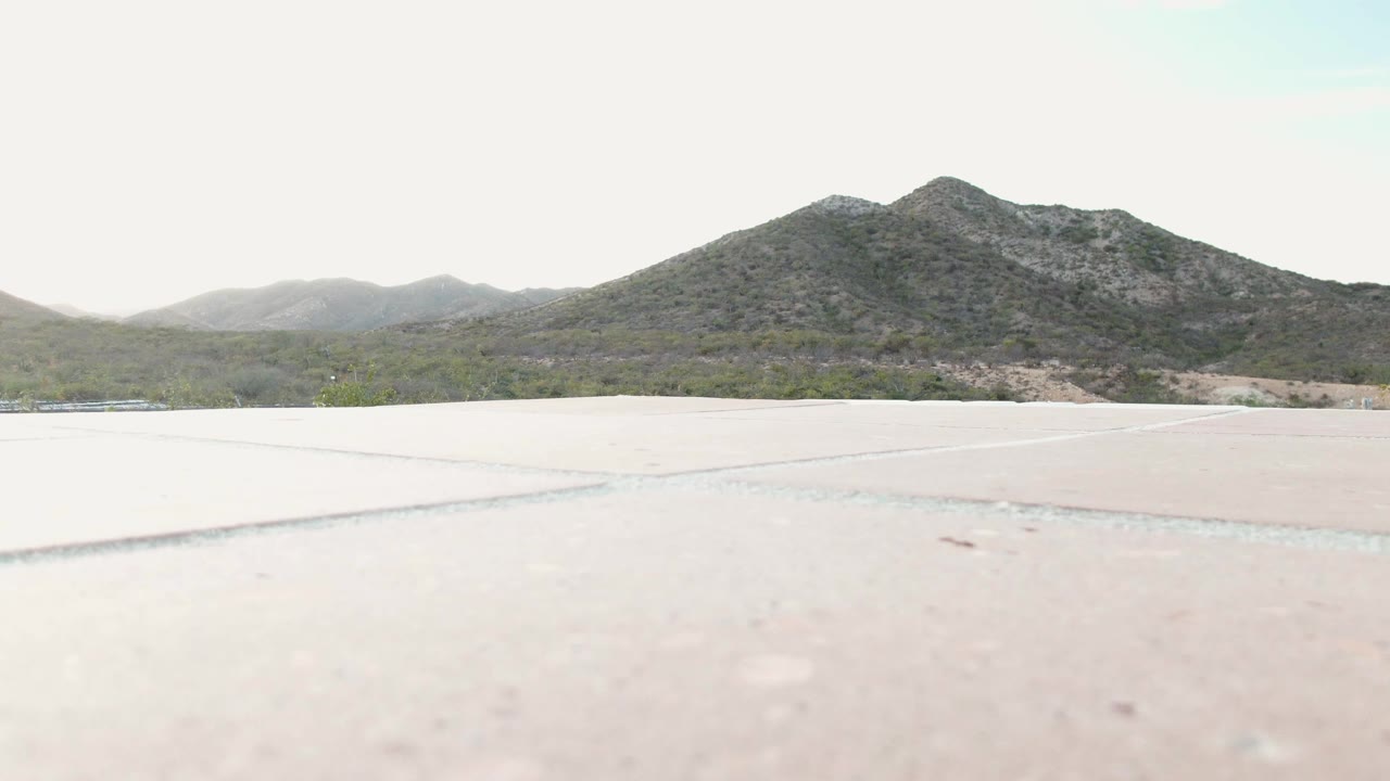 Cabo Pulmo, Baja California Sur frame showing a pale concrete expanse, a thin band of desert vegetation, and layered hills under bright haze, with no visible people.