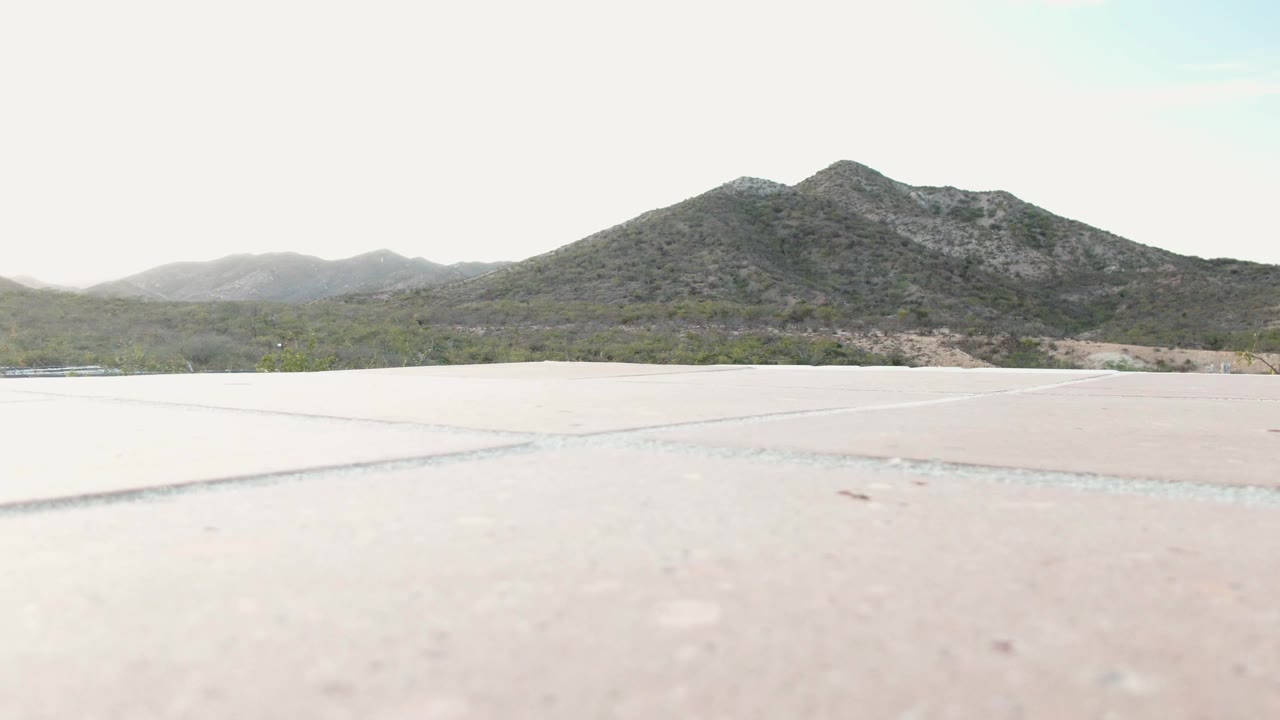 Low-angle view across pale concrete toward scrub vegetation and rocky mountains in Cabo Pulmo, Baja California Sur, Mexico, with no visible people in this opening frame.