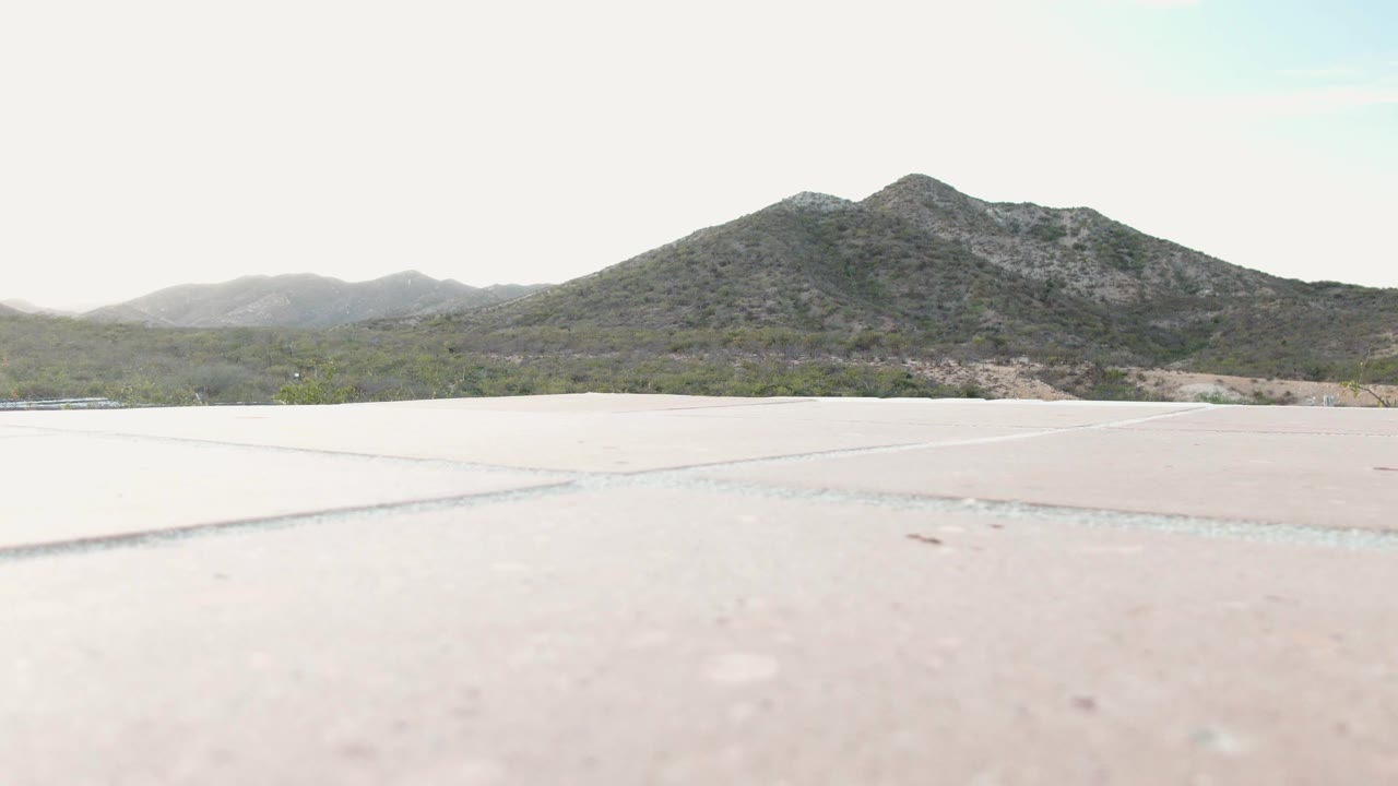 The frame shows a bright concrete or paved foreground, low desert vegetation across the horizon, and rocky mountains in the background near Cabo Pulmo, with no visible people.