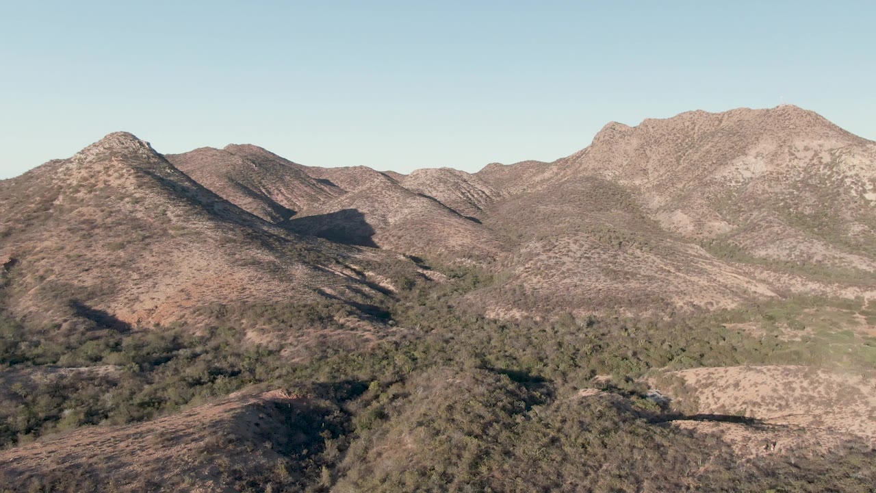 Opening frame of Cabo Pulmo, Baja California Sur, with brown rocky ridges, green vegetation in the valley, clear blue sky, and no visible people.