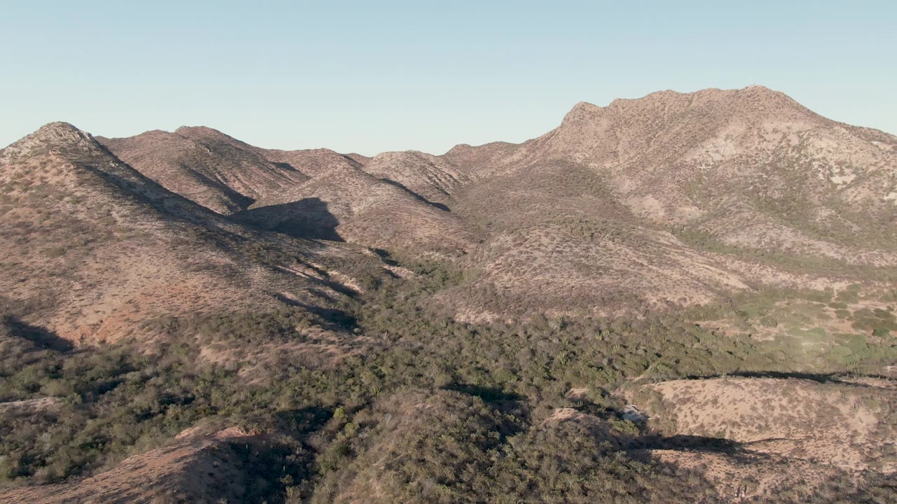 Later frame of the Cabo Pulmo aerial pan, with a dry hillside in the foreground, green vegetation through the valley, a broad rocky ridge behind it, and no visible people.