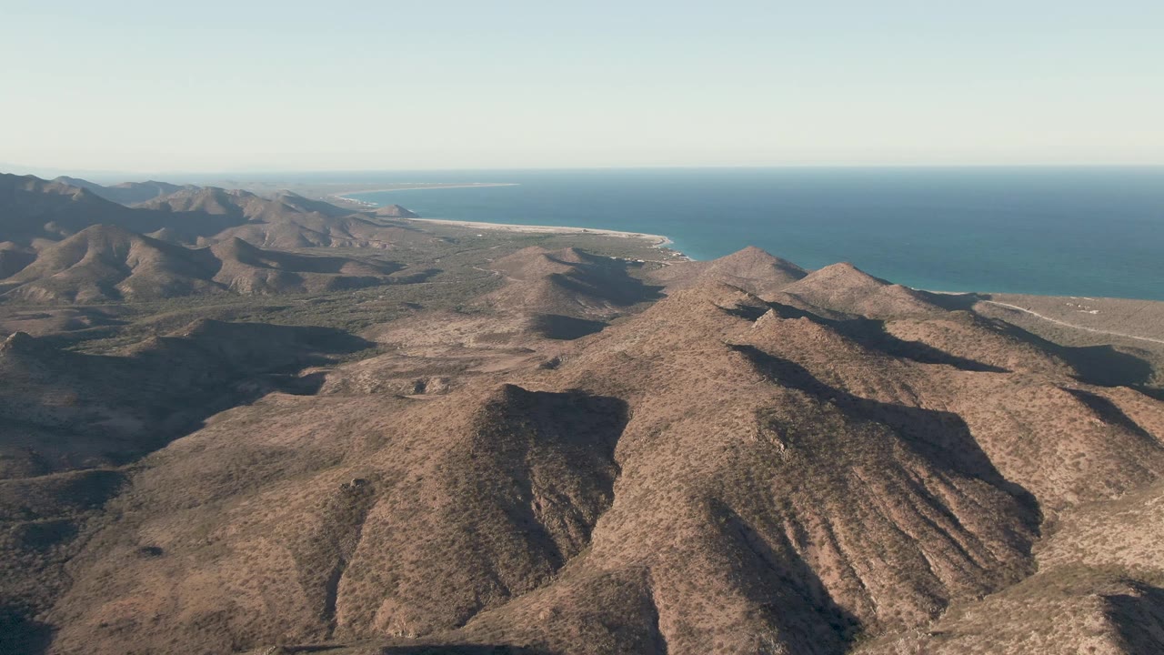 At 0:02, a centered arid ridge in Cabo Pulmo sits below a wide blue ocean band and pale sky, with no visible people or vehicles.
