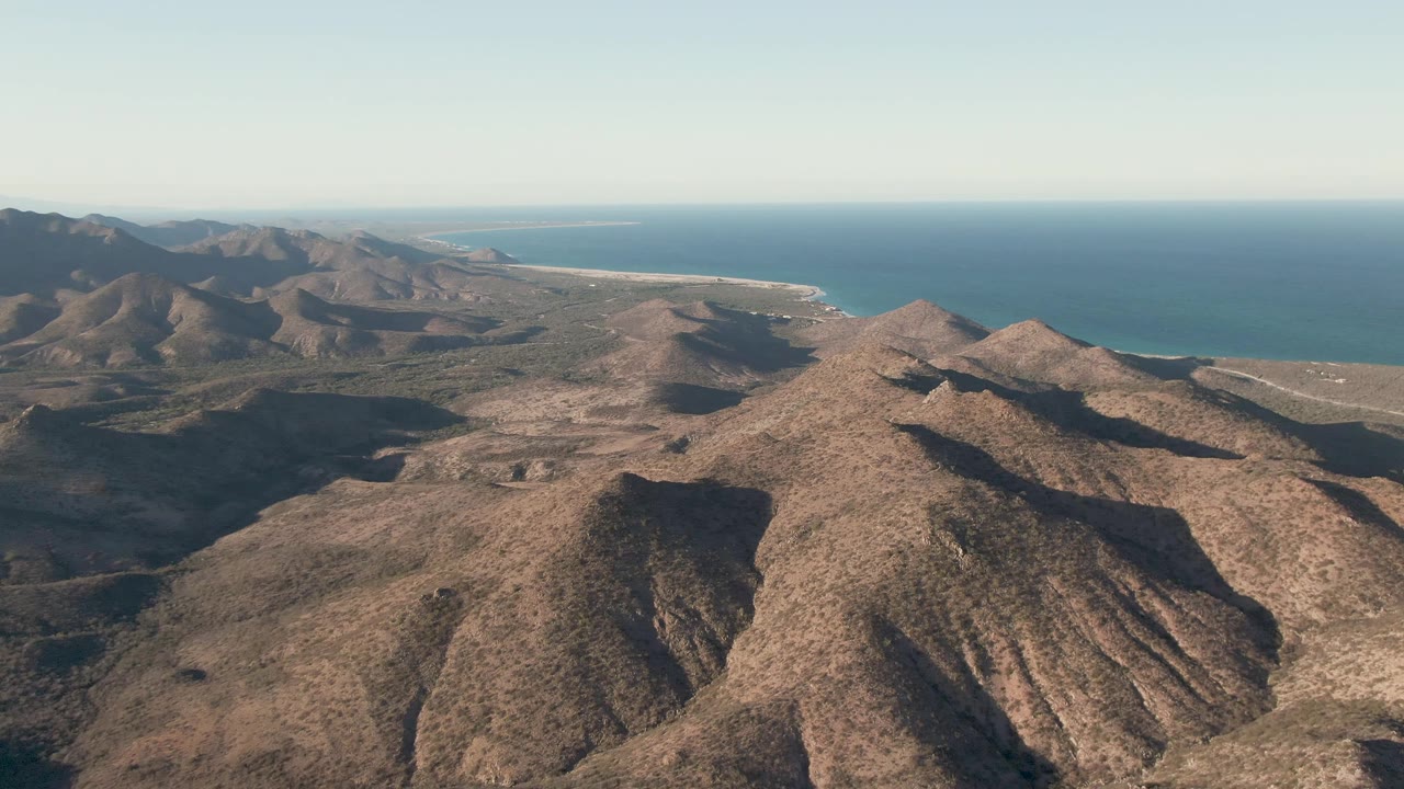 At 0:03, the aerial view over Cabo Pulmo shows layered brown hills and long shadows with the sea and coastline stretching across the background, with no visible people.