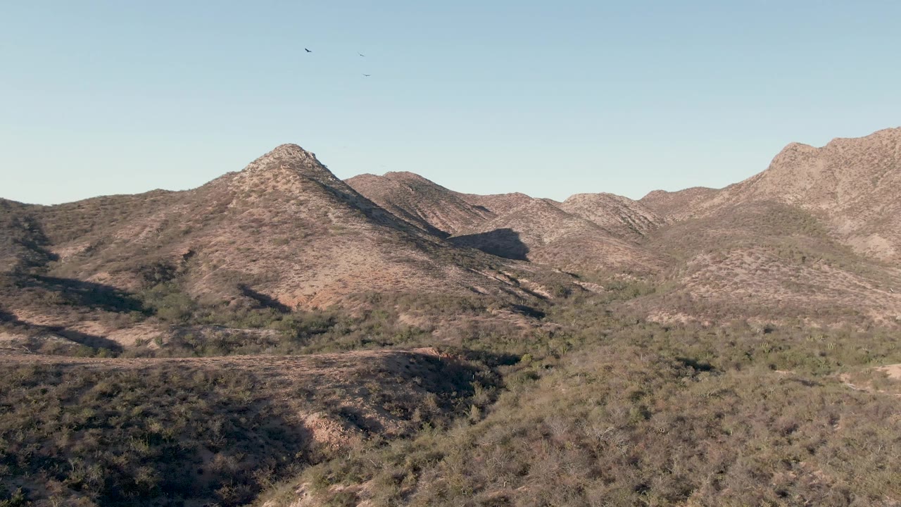 Wide elevated view of arid mountain ridges in Cabo Pulmo, Baja California Sur, with sparse green vegetation below a clear sky and no visible people.