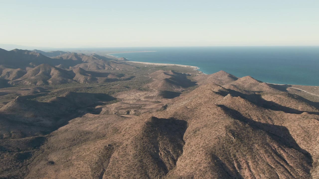 An aerial view across Cabo Pulmo's arid hills, with layered ridges leading toward a distant coastline and no visible people.