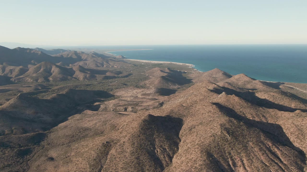 A high aerial frame in Cabo Pulmo shows rocky desert hills, a faint winding road through the terrain, ocean beyond the ridges, and no visible people.