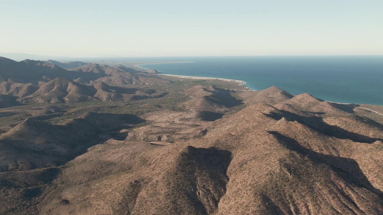 A later aerial frame over Cabo Pulmo shows road traces across the dry mountain basin, with the coastline and blue sea stretching across the background and no visible people.
