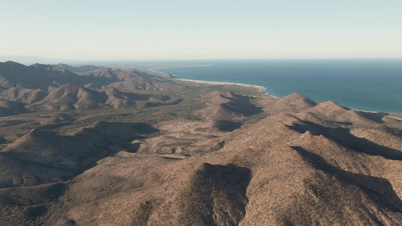 Opening aerial frame over Cabo Pulmo, Baja California Sur, showing rocky desert ridges in front of a blue ocean coastline with no visible people.