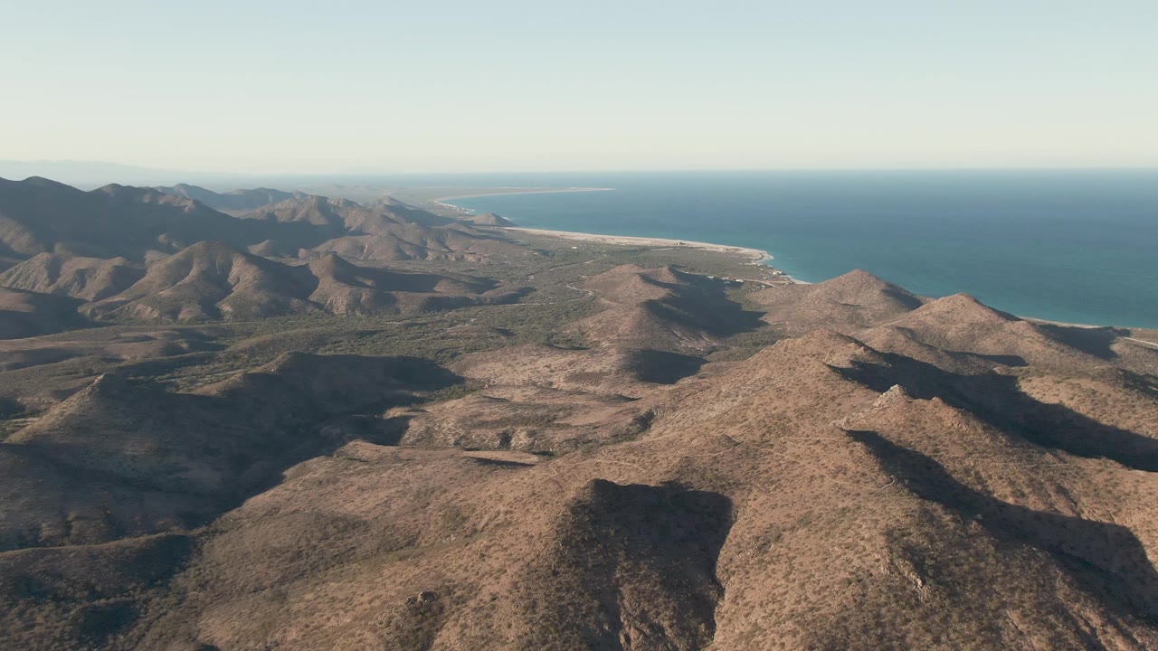 Early aerial frame over Cabo Pulmo with sunlit tan ridges leading toward a narrow beach, blue water, and distant mountains, with no visible people.