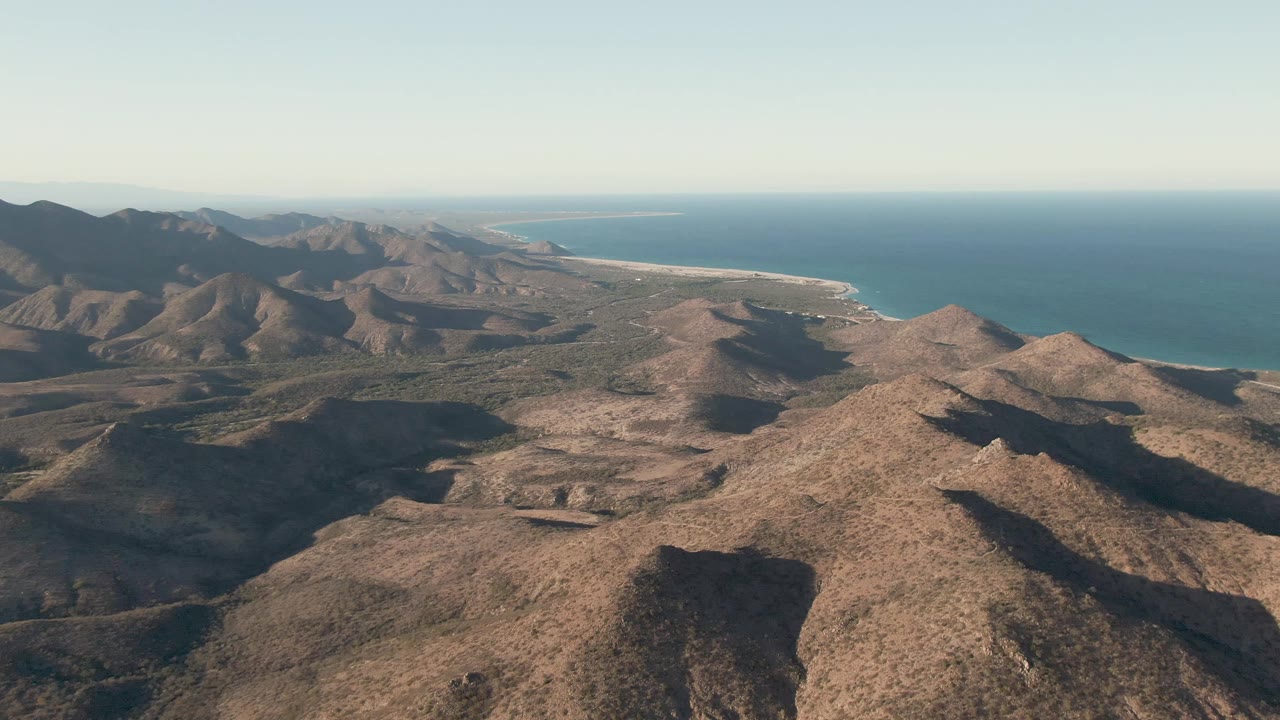 Aerial frame in Cabo Pulmo showing rugged brown hills, shadowed valleys, a pale coastal strip, and open blue ocean, with no visible people.