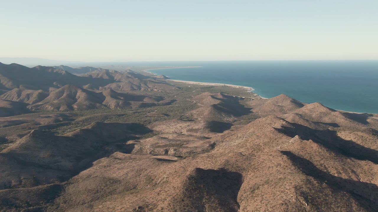Wide drone view of rocky desert mountains beside a blue ocean coastline.