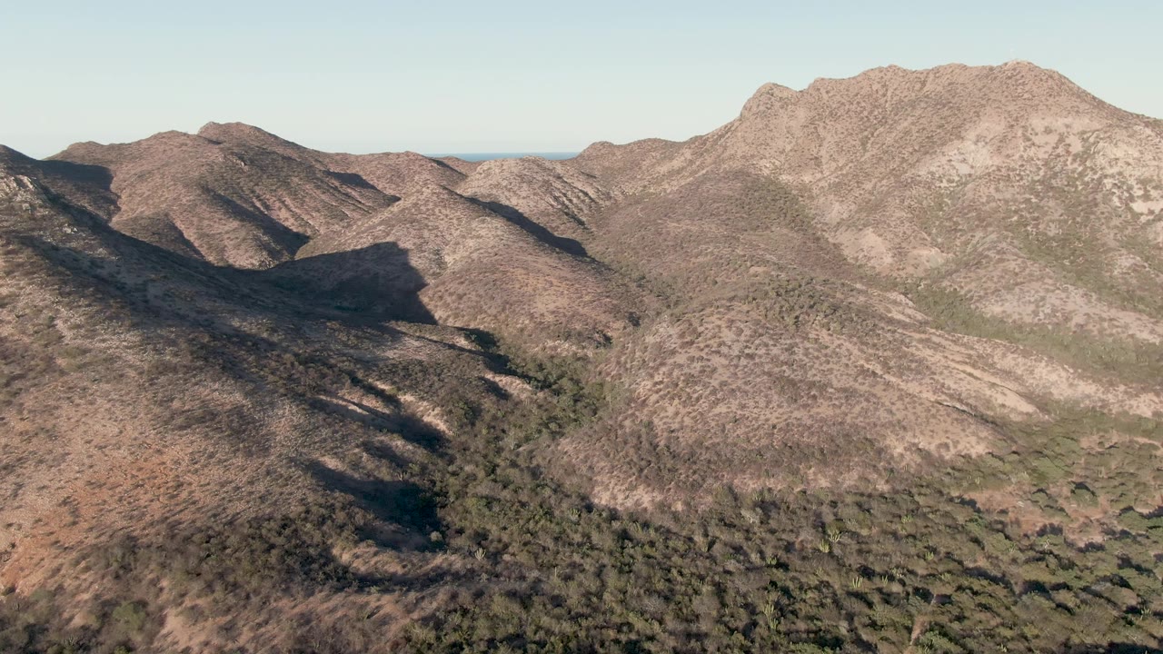 Aerial view over Cabo Pulmo, Baja California Sur, showing dry brown mountain ridges, dark valley shadows, sparse green vegetation, distant ocean horizon, and no visible people.