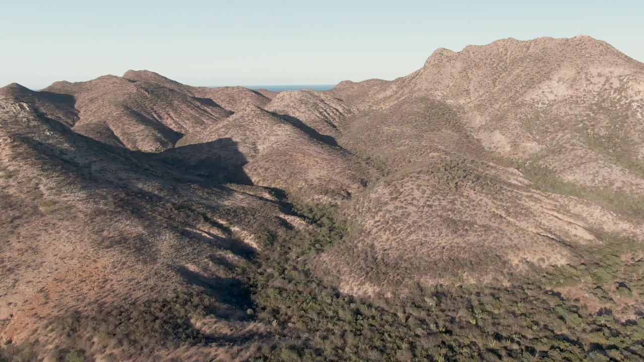 Aerial frame from Cabo Pulmo, Baja California Sur, with rocky brown slopes across the foreground, a darker central valley, a thin ocean line in the distance, and no visible people.