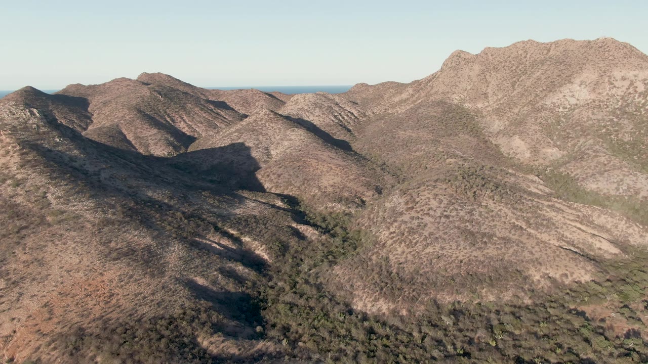 Cabo Pulmo aerial view with a sunlit high ridge on the right, lower brown hills to the left, green vegetation in the valley floor, distant ocean, and no visible people.