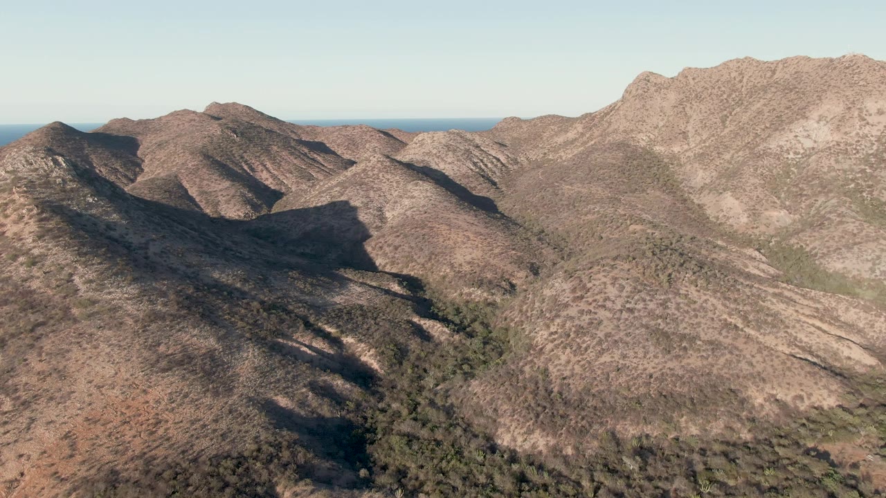 Aerial mountain landscape with broad dry slopes, dark gullies, and ocean horizon in back.