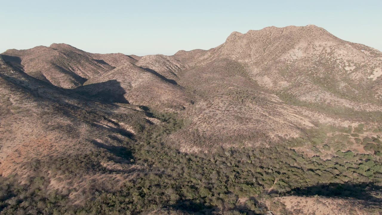 Cabo Pulmo aerial frame with the left hillside in deeper shadow, sunlit brown slopes to the right, green vegetation low in frame, and no visible people.