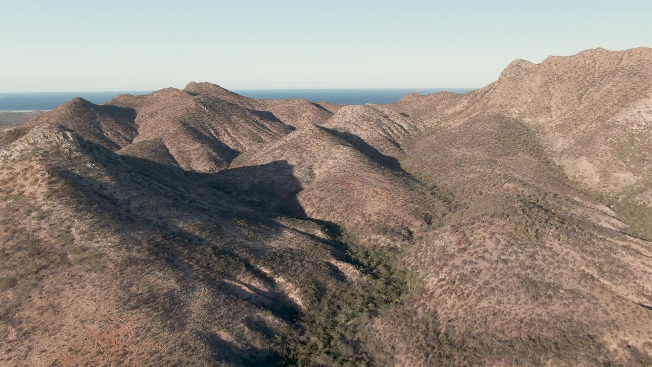 Aerial frame over Cabo Pulmo showing dry brown mountain ridges, deep valley shadows, a thin blue ocean horizon, clear sky, and no visible people.