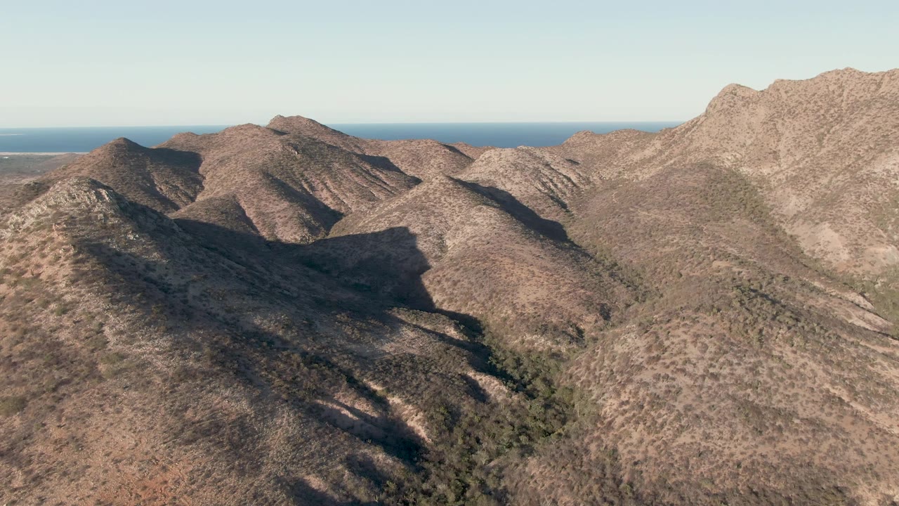 A second aerial frame in Cabo Pulmo shows overlapping dry ridges, a broad dark shadow across the central slope, blue ocean in the distance, and no visible people.