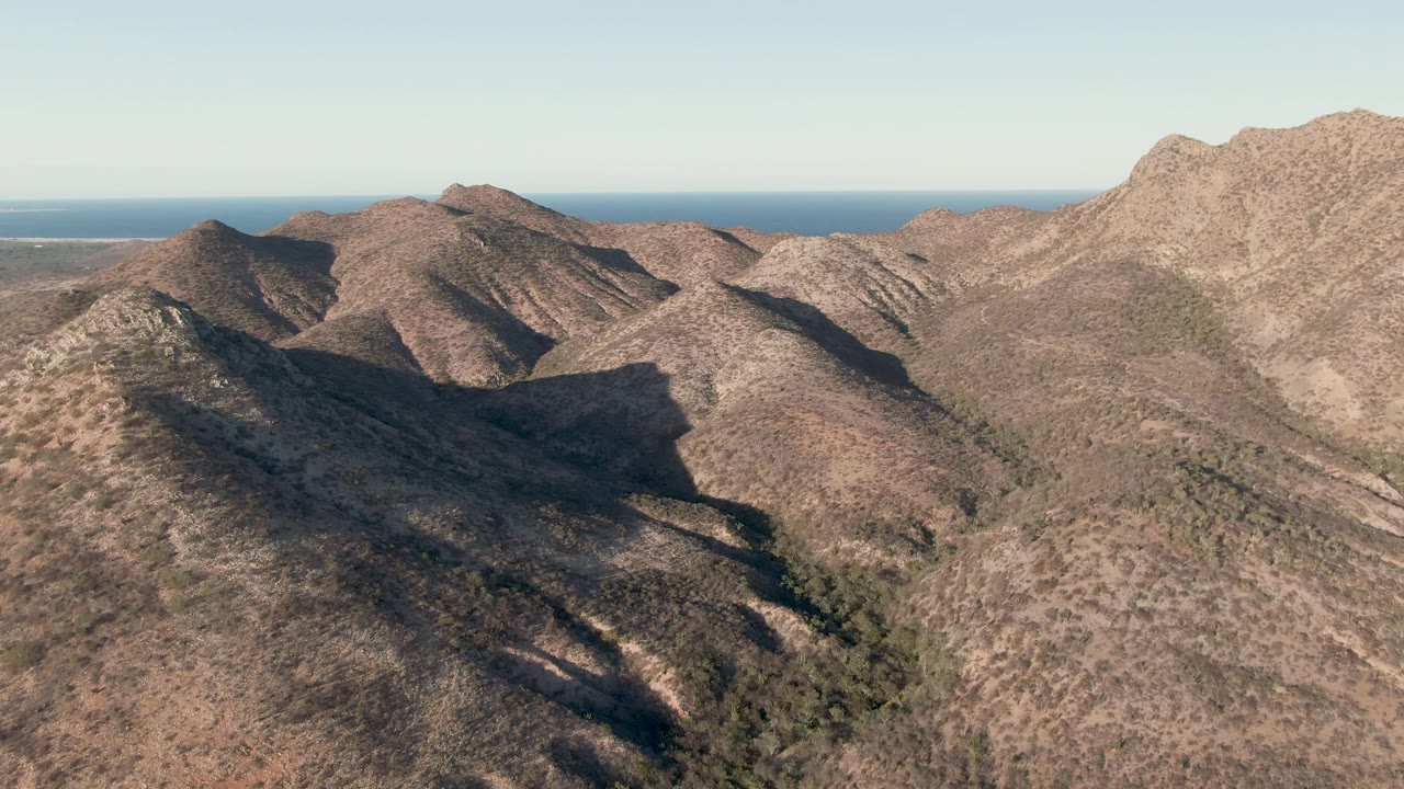 Aerial view near Cabo Pulmo with sunlit brown hills, darker valleys, rugged ridgelines, a level ocean horizon, and no visible people.