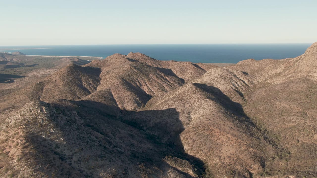 Aerial frame of layered arid hills near Cabo Pulmo with broad ridge shadows, sparse vegetation, ocean in the background, and no visible people.