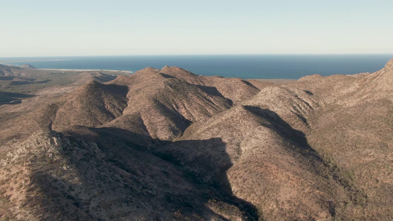 High aerial view across sunlit rocky ridges in Cabo Pulmo, with the ocean line stretching behind the hills and no visible people.