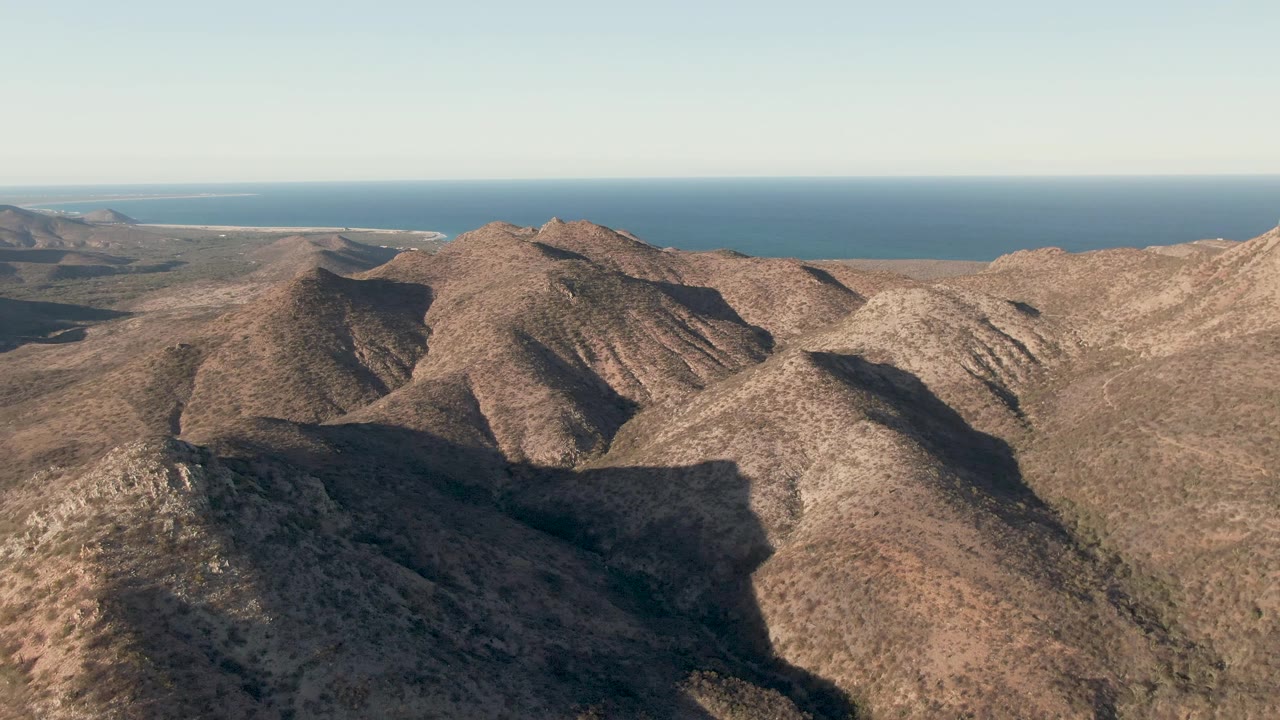 Mid-clip aerial composition over dry Cabo Pulmo slopes, with a large shadowed ridge in the foreground, distant ocean, and no visible people.