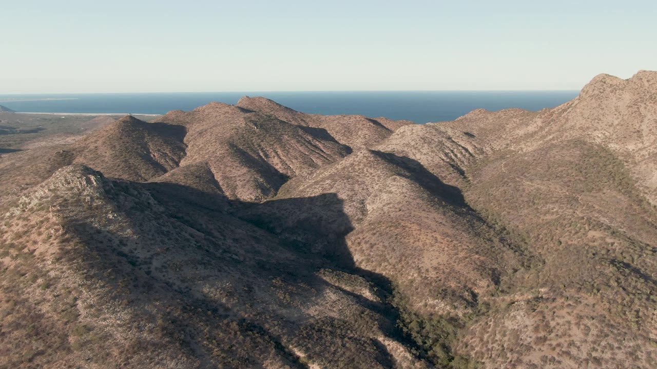 Rocky brown ridges in Cabo Pulmo stretch across the frame, with a darker central valley shadow, thin ocean band, pale sky, and no visible people.
