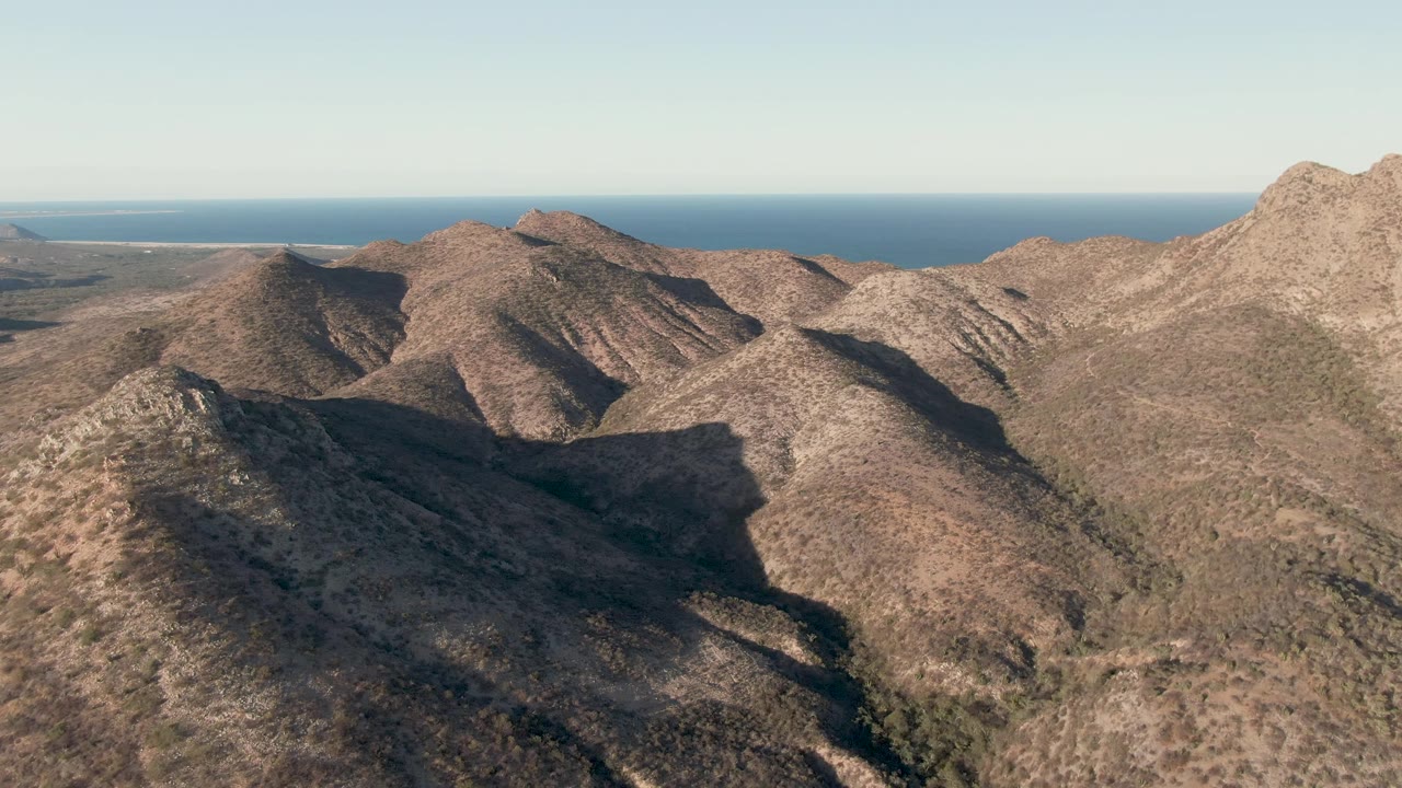 Sunlit and shadowed desert ridges in Cabo Pulmo fill the lower frame, with sparse vegetation, a blue ocean strip beyond the hills, and no visible people.