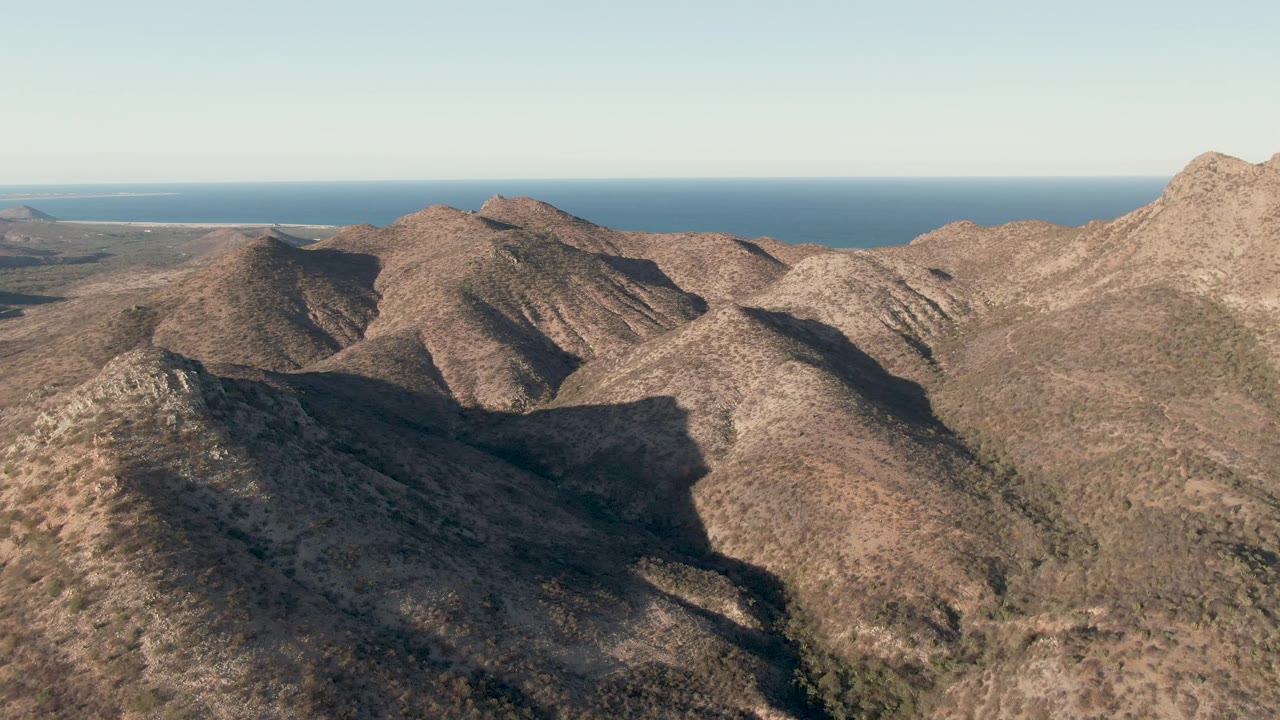 Dry ridgelines in Cabo Pulmo angle across the foreground and midground, with strong hillside shadows, open scrub terrain, distant ocean, and no visible people.