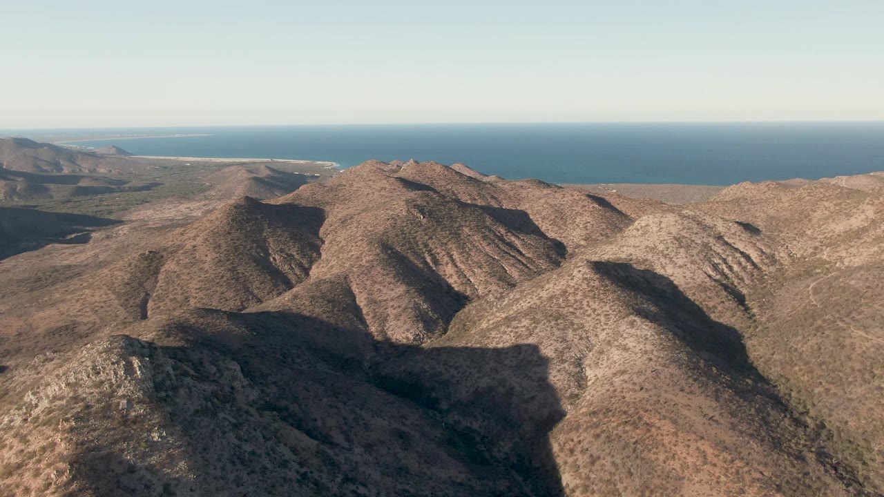 Opening frame over Cabo Pulmo mountain ridges, with brown rocky slopes in front, blue ocean beyond, clear sky above, and no visible people.