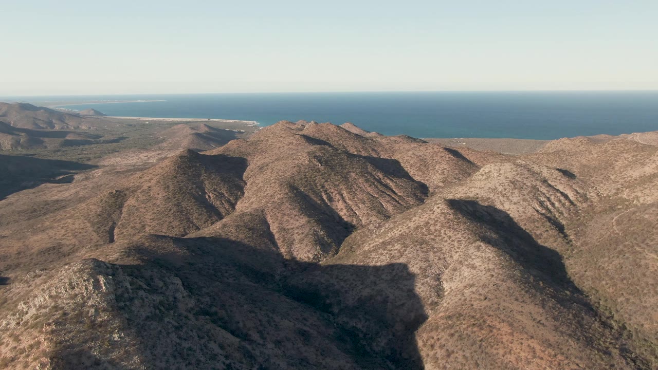 Second frame of the Cabo Pulmo aerial, showing the central ridge more prominently with dark valley shadow at lower center, distant coastline, and no visible people.