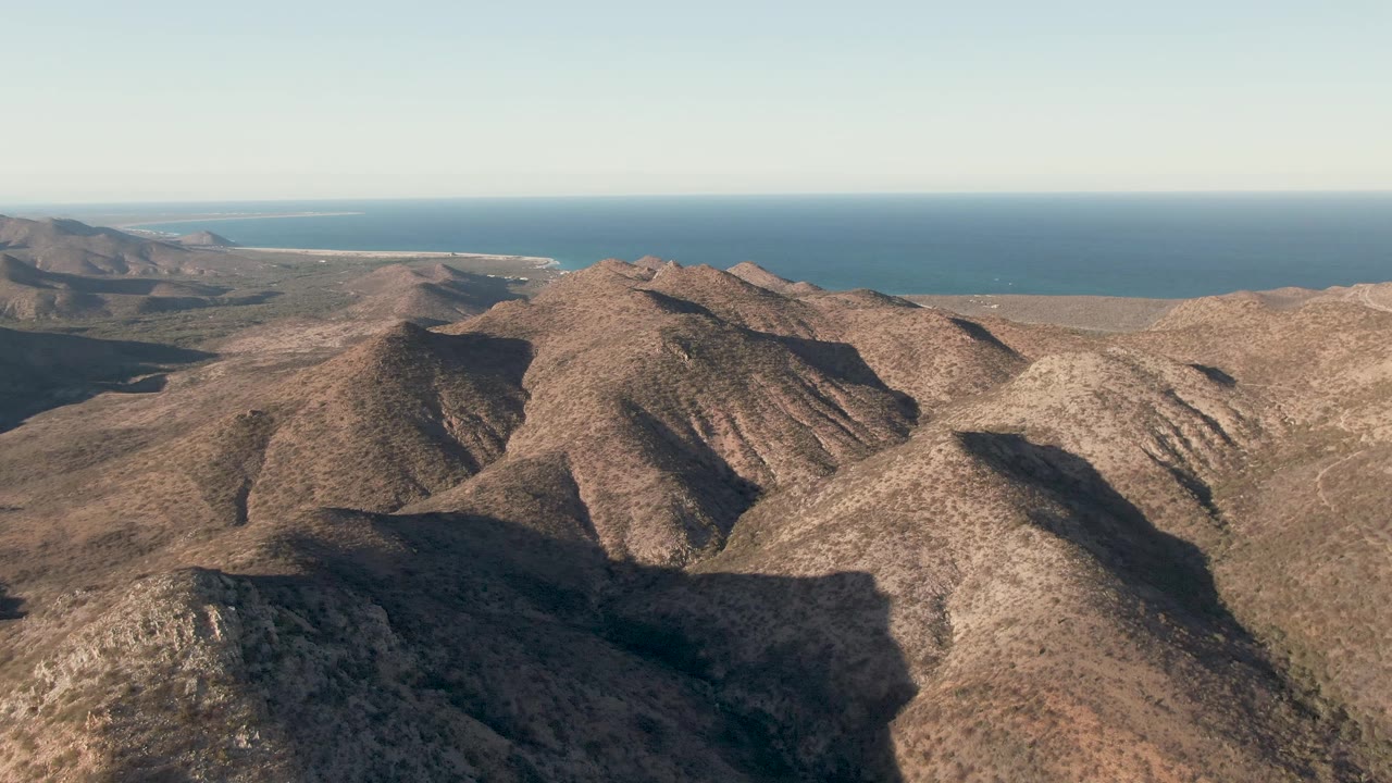 Third frame over Cabo Pulmo ridgelines, with the foreground slope shifted slightly across the frame, a long blue ocean band in the background, and no visible people.