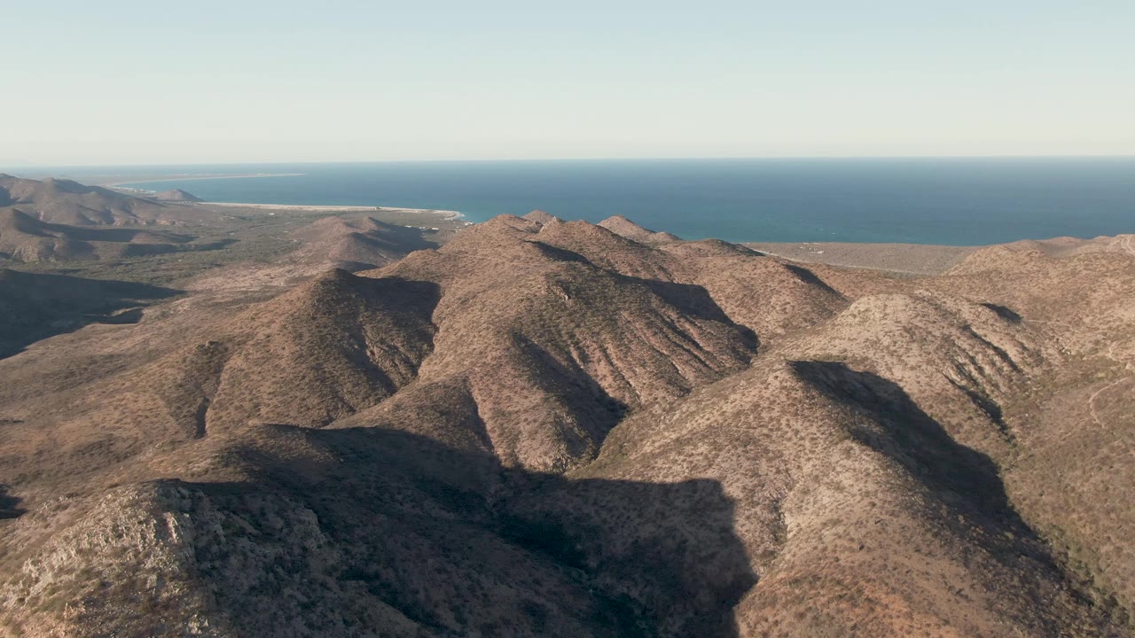 Fourth frame in Cabo Pulmo with broad arid ridges across the lower frame, stronger shadow in the valleys, blue ocean at the horizon, and no visible people.