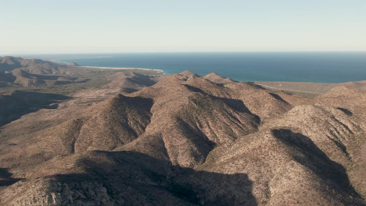 Second frame shows Cabo Pulmo mountain ridges from above, with a broad shadowed valley, dry brown slopes, a narrow coastline, blue ocean, and no visible people.