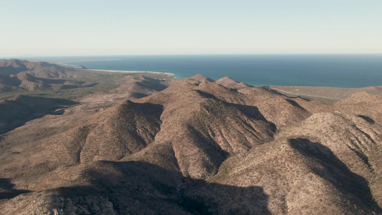 Aerial view in Cabo Pulmo with rocky foreground hills, overlapping mountain ridges, deep shadows on the left slope, calm ocean in the distance, and no visible people.