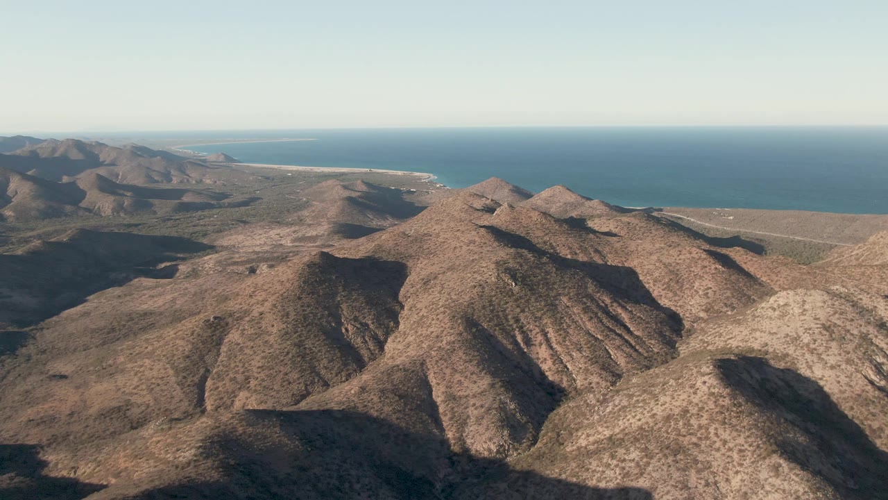 Aerial view over Cabo Pulmo, Baja California Sur, with sunlit brown ridges in front, distant blue ocean, clear sky, and no visible people.