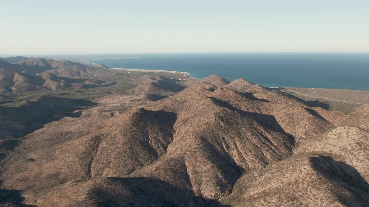 Aerial view in Cabo Pulmo, Baja California Sur, with a rounded foreground hill, jagged brown ridge forms, distant water, and no visible people.