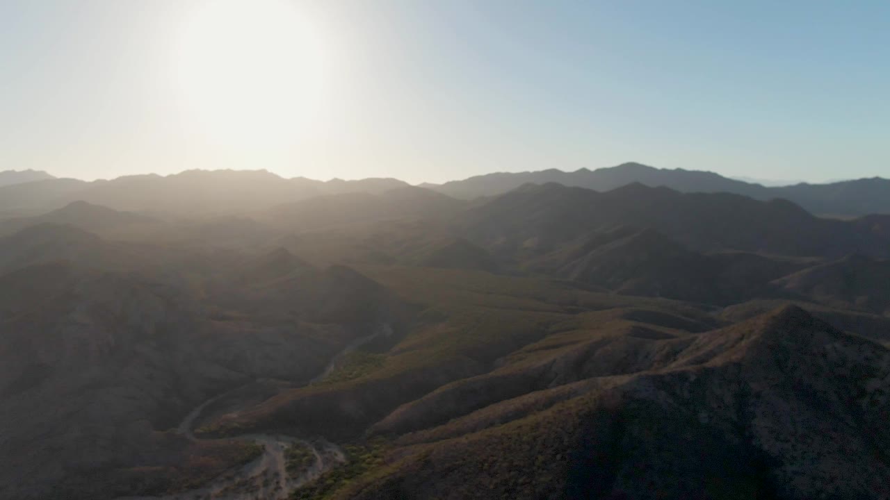 A backlit aerial view of Cabo Pulmo hills shows layered mountain ridges, a winding dirt road through the valley, scrub-covered slopes, and no visible people.