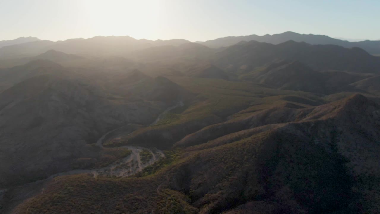 The aerial view looks across shadowed Cabo Pulmo hills under a bright low sun, with a winding road tracing the valley floor and no visible people.