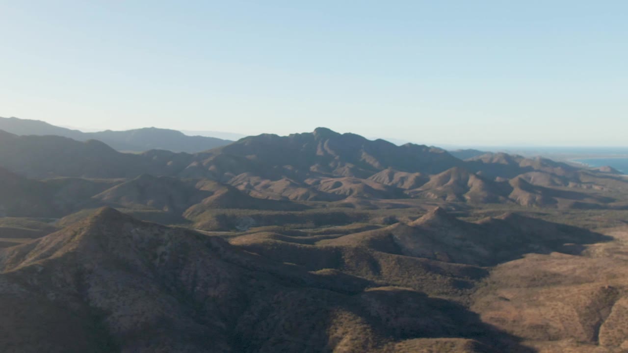 A wide aerial view shows Cabo Pulmo's dry valley floor, rugged mountain ridges, distant haze, and clear sky, with no visible people.