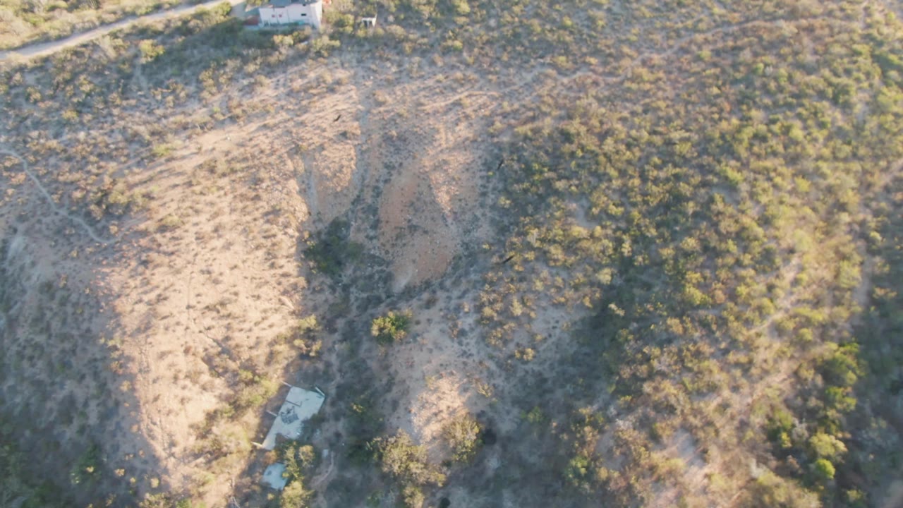 Aerial view in Cabo Pulmo, Baja California Sur with scrub-covered slopes, pale eroded ground, a small light-colored structure near the lower right, and no visible people.