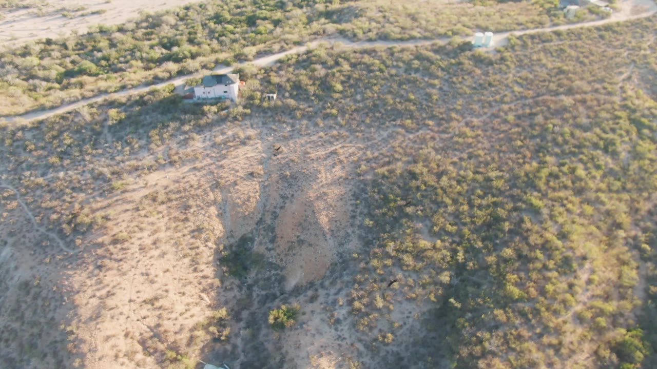 High aerial frame over Cabo Pulmo, Baja California Sur showing a dirt road along the upper edge, dry slopes, scattered green shrubs, a small structure, and no visible people.