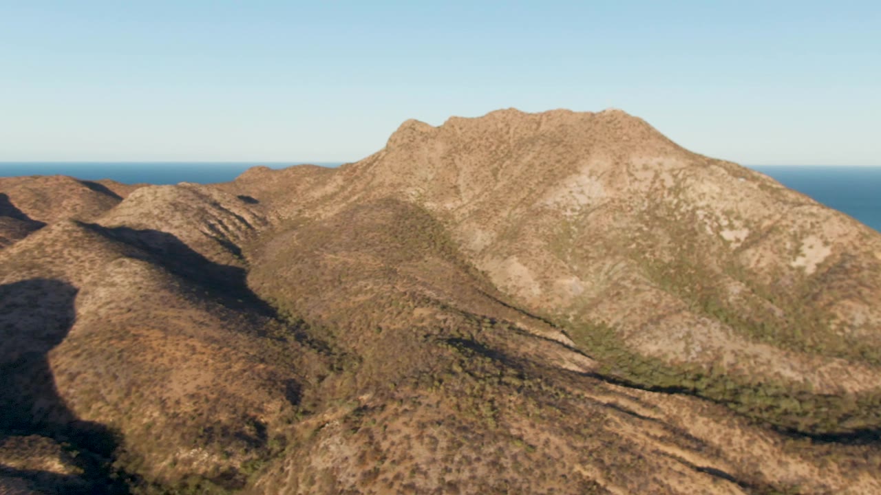 Wide aerial frame of Cabo Pulmo, Baja California Sur, showing a sunlit mountain ridge, dry hills, blue ocean horizon, clear sky, and no visible people.