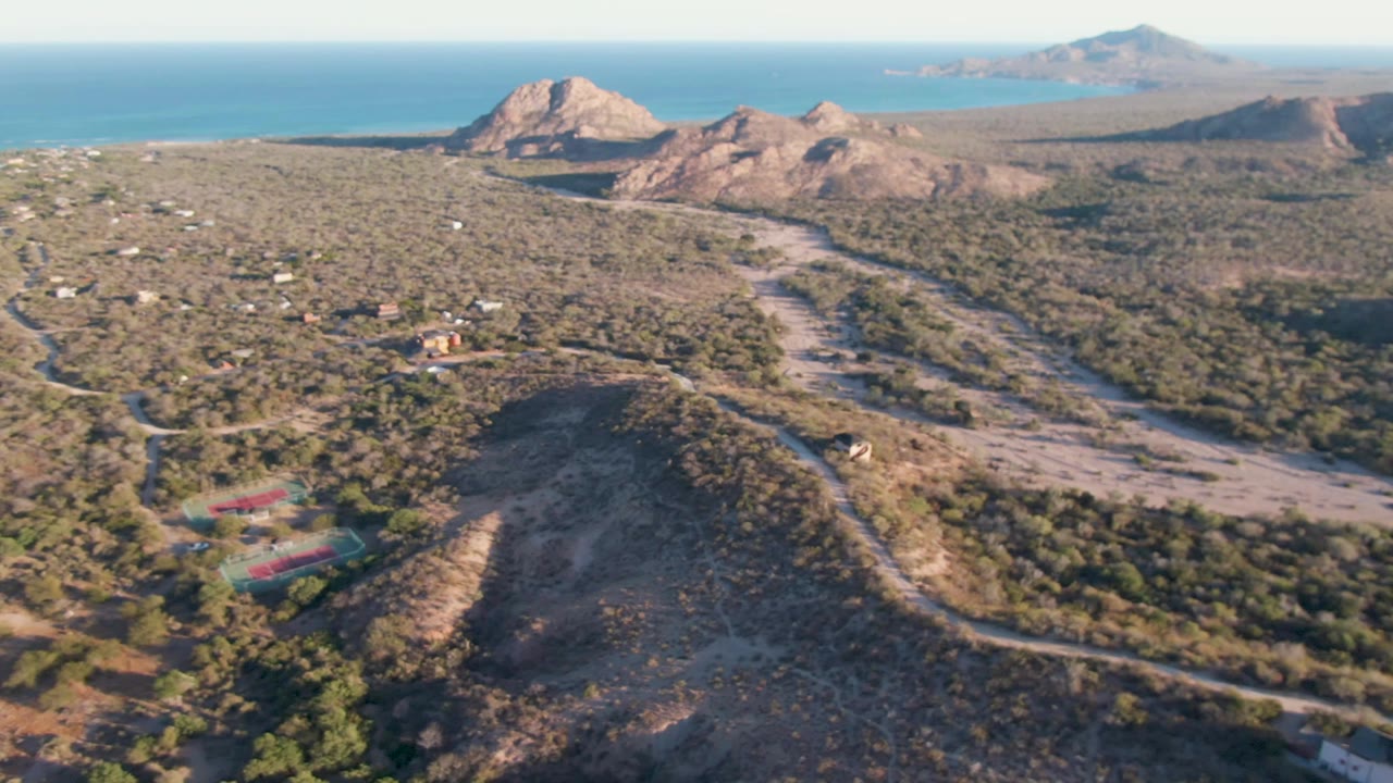 High aerial frame over Cabo Pulmo showing a dry valley, small homes, pale dirt roads, mountain ridges, ocean in the distance, and no visible people.
