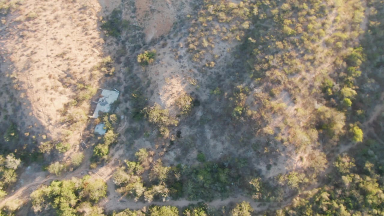 A steep aerial view over a dry hillside in Cabo Pulmo, Baja California Sur, with scattered shrubs, dirt tracks, a small light-roofed building near the lower left, and no visible people.