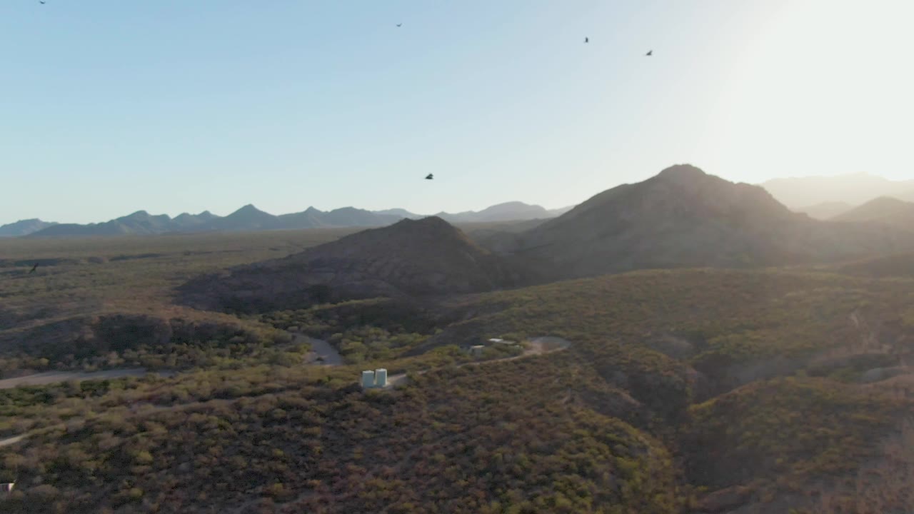 Cabo Pulmo desert hills stretch toward layered mountains as bright sun enters from the upper right, with birds overhead, small structures below, and no visible people.